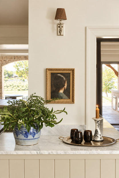 A framed portrait print hanging on a light gray wall in a home interior, with a marble countertop below holding a potted plant, dark cups, and a candle holder. The portrait depicts a person from behind wearing a dark top and a gold-toned frame.