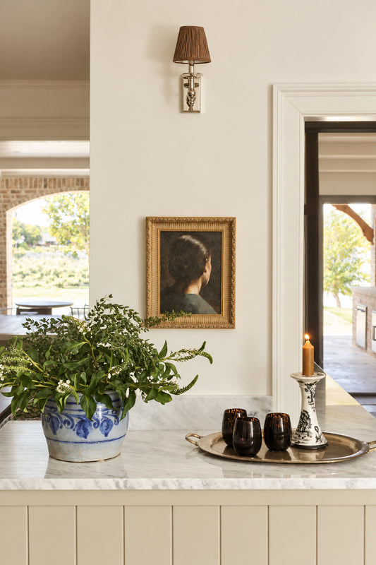 A framed portrait print hanging on a light gray wall in a home interior, with a marble countertop below holding a potted plant, dark cups, and a candle holder. The portrait depicts a person from behind wearing a dark top and a gold-toned frame.