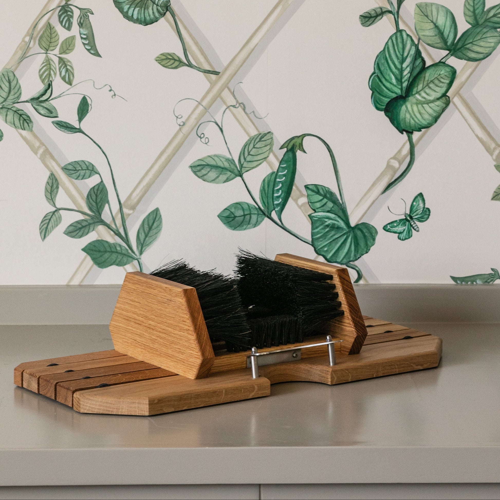 Wooden boot cleaner with black brushes against a decorative wall with green leaves.