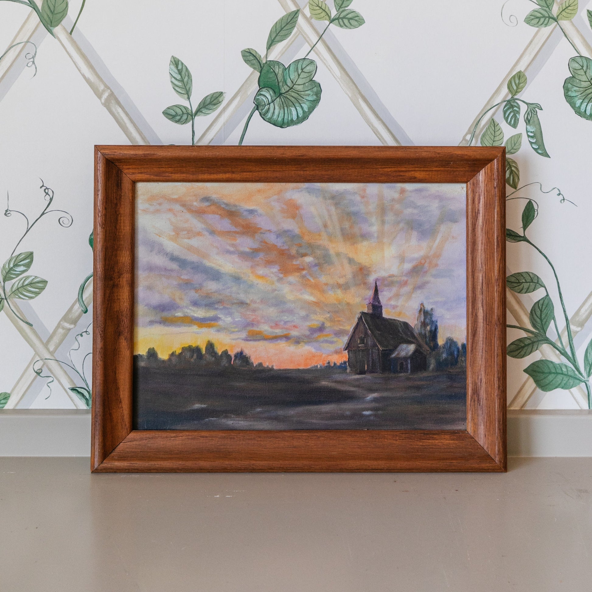 Framed painting of a sunset over a church with decorative wallpaper in the background