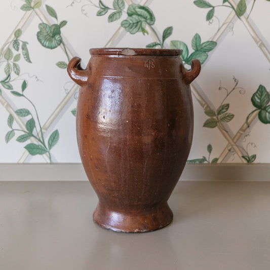 Brown ceramic jar on a gray surface with a decorative green and white patterned wall in the background