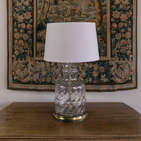 Clear glass lamp with white shade on a wooden table in front of an ornate tapestry.