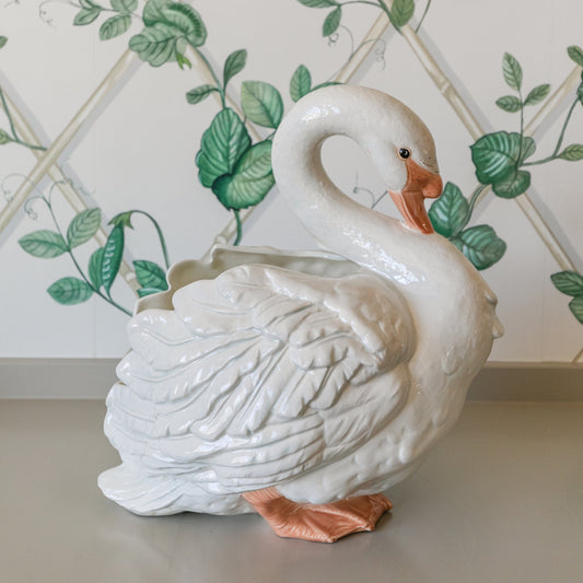 White ceramic swan planter on a surface with decorative green leaf patterned wallpaper.