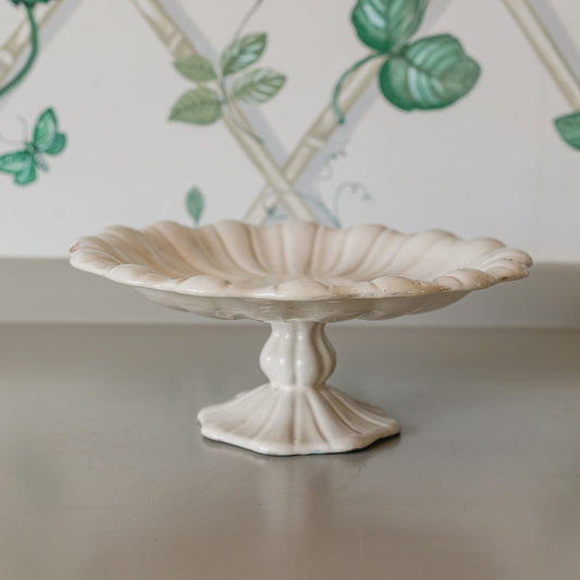 Decorative white pedestal dish on a neutral surface with a decorative wall in the background