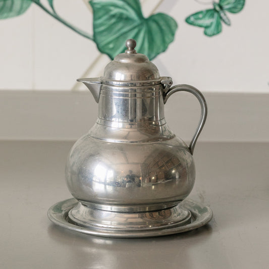 Silver pitcher on a reflective surface with a decorative wall in the background