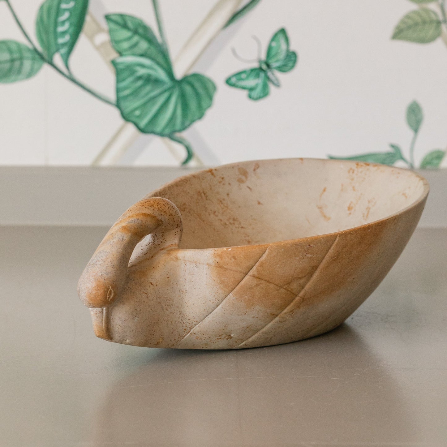 Stone planter shaped like a swan on a surface with a decorative tile background featuring green leaves and a butterfly.