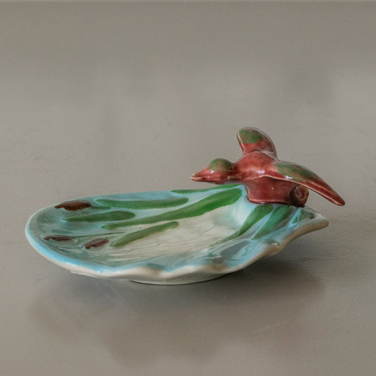 Ceramic duck on a leaf-shaped plate on a gray surface with a floral-patterned curtain in the background.